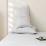 Two Linenly Luxe Cotton Sateen Pillowcases in silver are stacked on a neatly made bed with white bedding. A tray with a cup, saucer, spoon, and book rests in the corner as sunlight casts soft shadows on the wall.