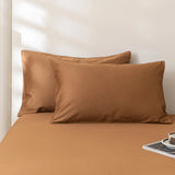 Two Luxe Cotton Sateen Pillowcases in Terracotta by Linenly are set against a matching bedsheet on a neatly made bed. Sunlight casts soft shadows behind, with a book and cup placed on the lower right bedside corner.