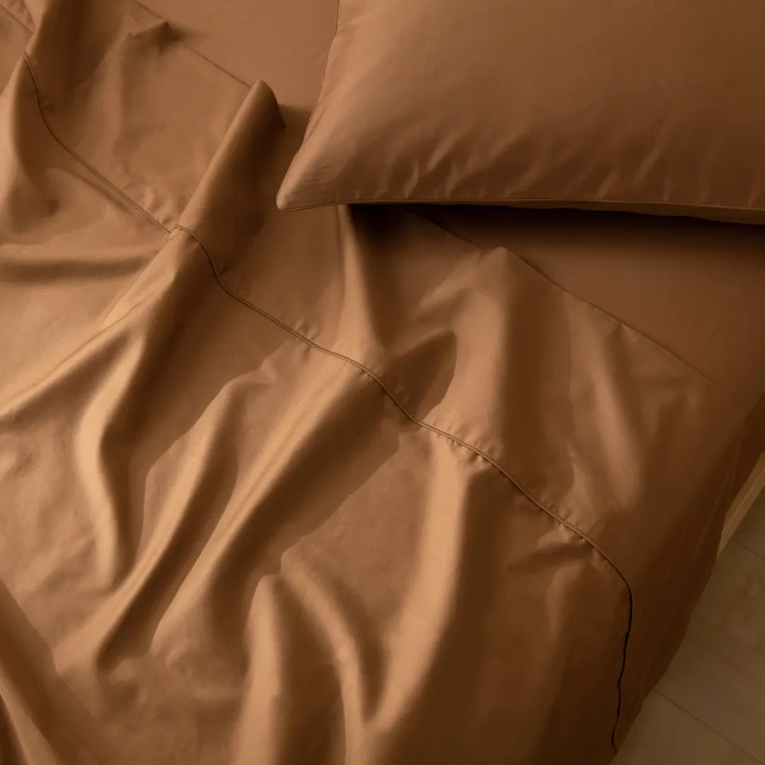 A close-up shows a neatly made bed with brown sheets and Linenly’s Luxe Cotton Sateen Pillowcase Set in Terracotta. The smooth, soft folds create a cozy, inviting look.