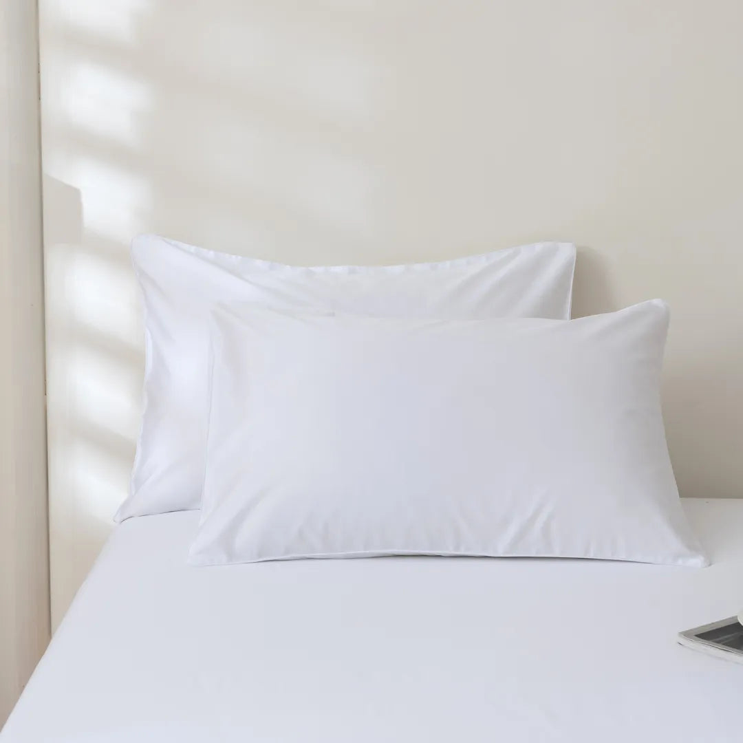Two pillows with the Linenly Luxe Cotton Sateen Pillowcase Set in white rest on a neatly made bed against a white wall, with sunlight casting striped shadows and a corner of a tablet visible on the bed.