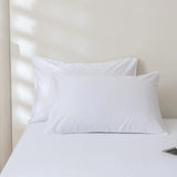 Two pillows with the Linenly Luxe Cotton Sateen Pillowcase Set in white rest on a neatly made bed against a white wall, with sunlight casting striped shadows and a corner of a tablet visible on the bed.