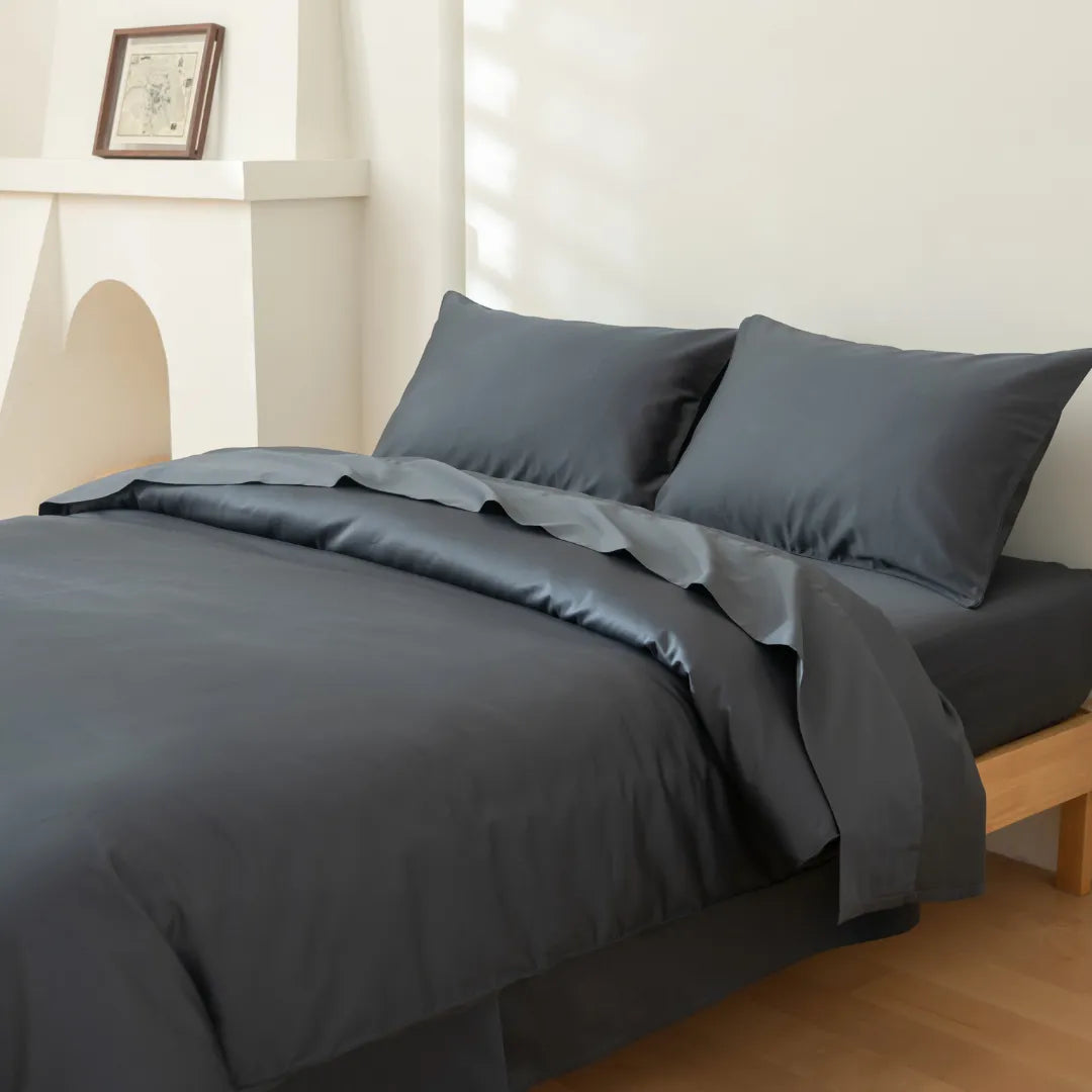 A minimalist room features a wooden bed dressed in Linenly's Luxe Cotton Sateen Quilt Cover in Charcoal, paired with a comforter and two pillows, set against white walls with a small mantel and framed picture.