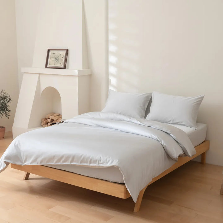 A neatly made bed with Linenly's Luxe Sateen Quilt Cover in Silver, made from white, 500 thread count long staple cotton bedding, in a serene bedroom with a minimalist aesthetic, featuring a decorative fireplace and natural light filtering through sheer curtains.