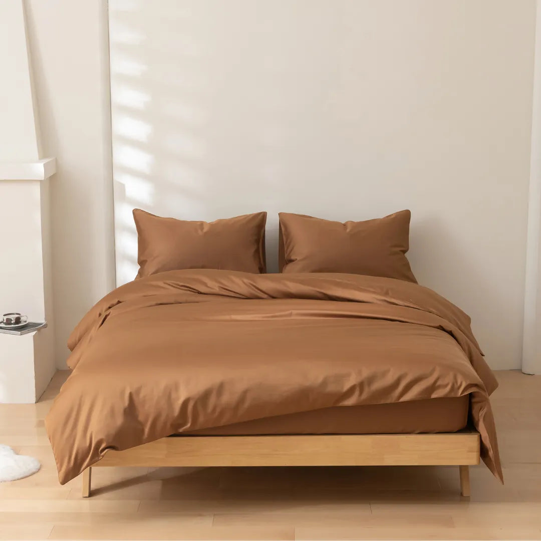A minimalist room with light wood floors features a neatly made bed dressed in Linenly’s Luxe Cotton Sateen Quilt Cover in Terracotta and two matching pillows. Sunlight casts soft shadows on the plain, light-colored wall.