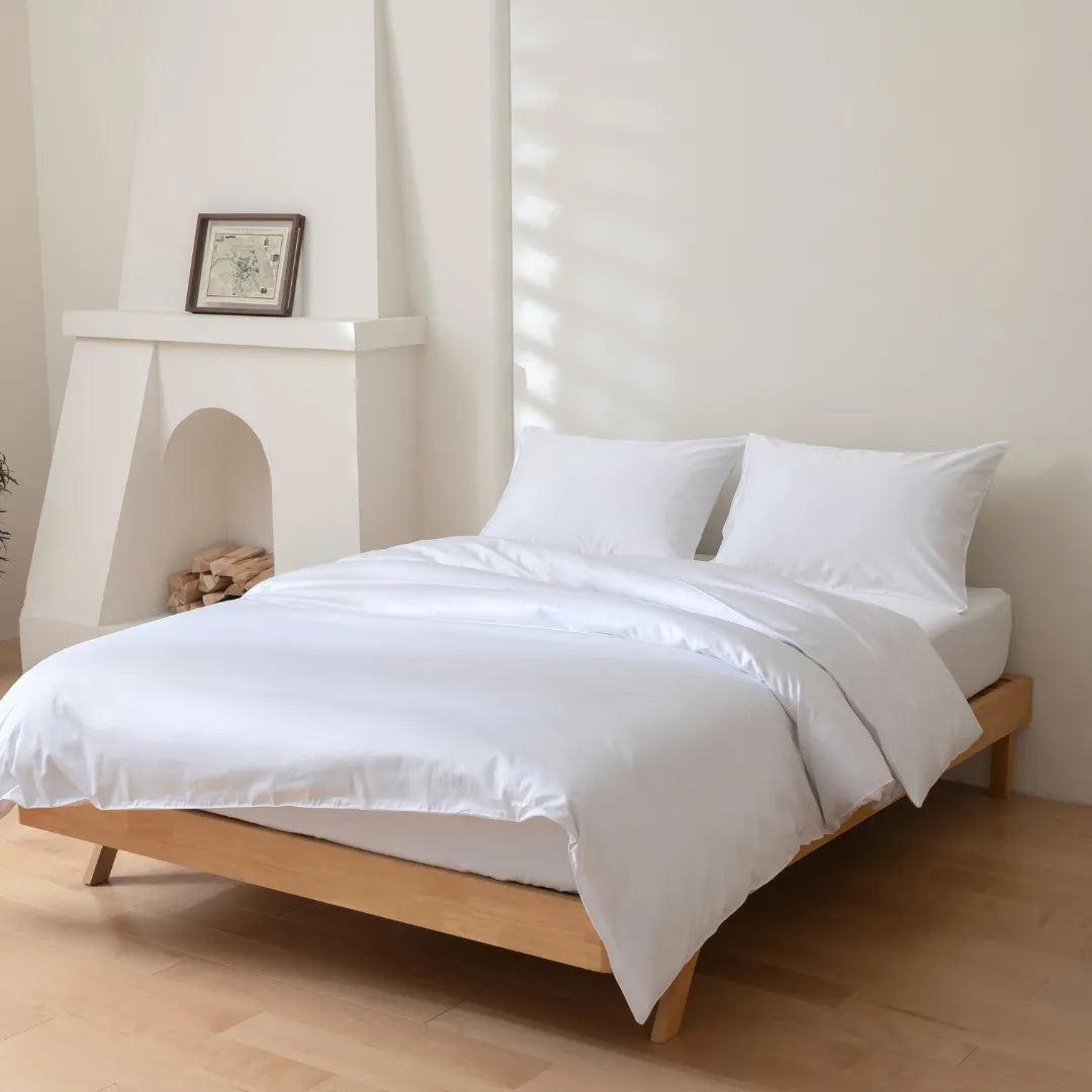 A neatly made bed with Linenly's Luxe Sateen Quilt Cover in white, 500 thread count sateen weave bedding in a bright minimalist bedroom, featuring a wooden platform bed frame and a decorative fireplace with logs.