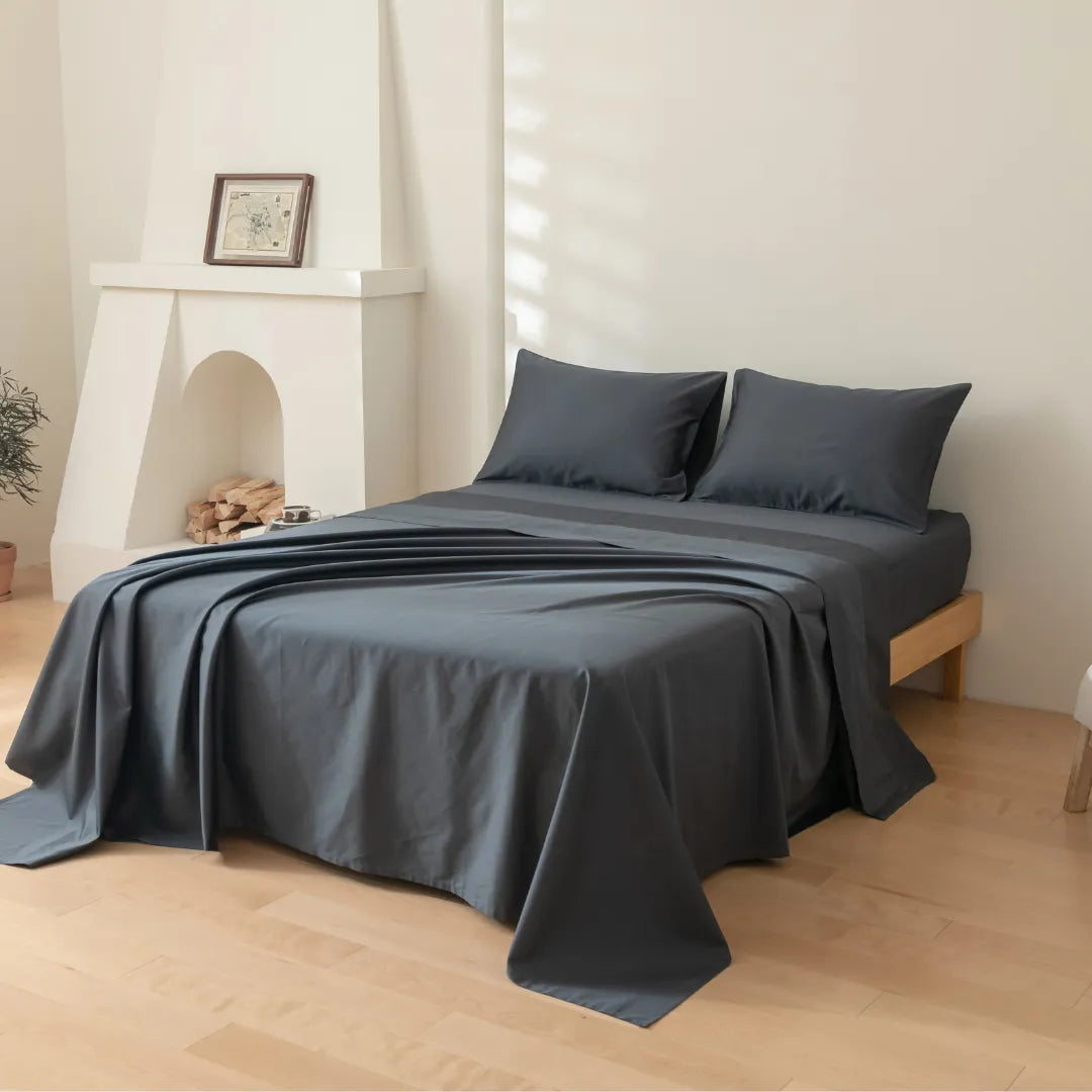 A minimalist room with light wooden floors features a neatly made bed dressed in Linenly’s Luxe Cotton Sateen Sheet Set in Charcoal, paired with two matching pillows. Soft natural light fills the space, and a framed picture rests on the mantel above the white fireplace.