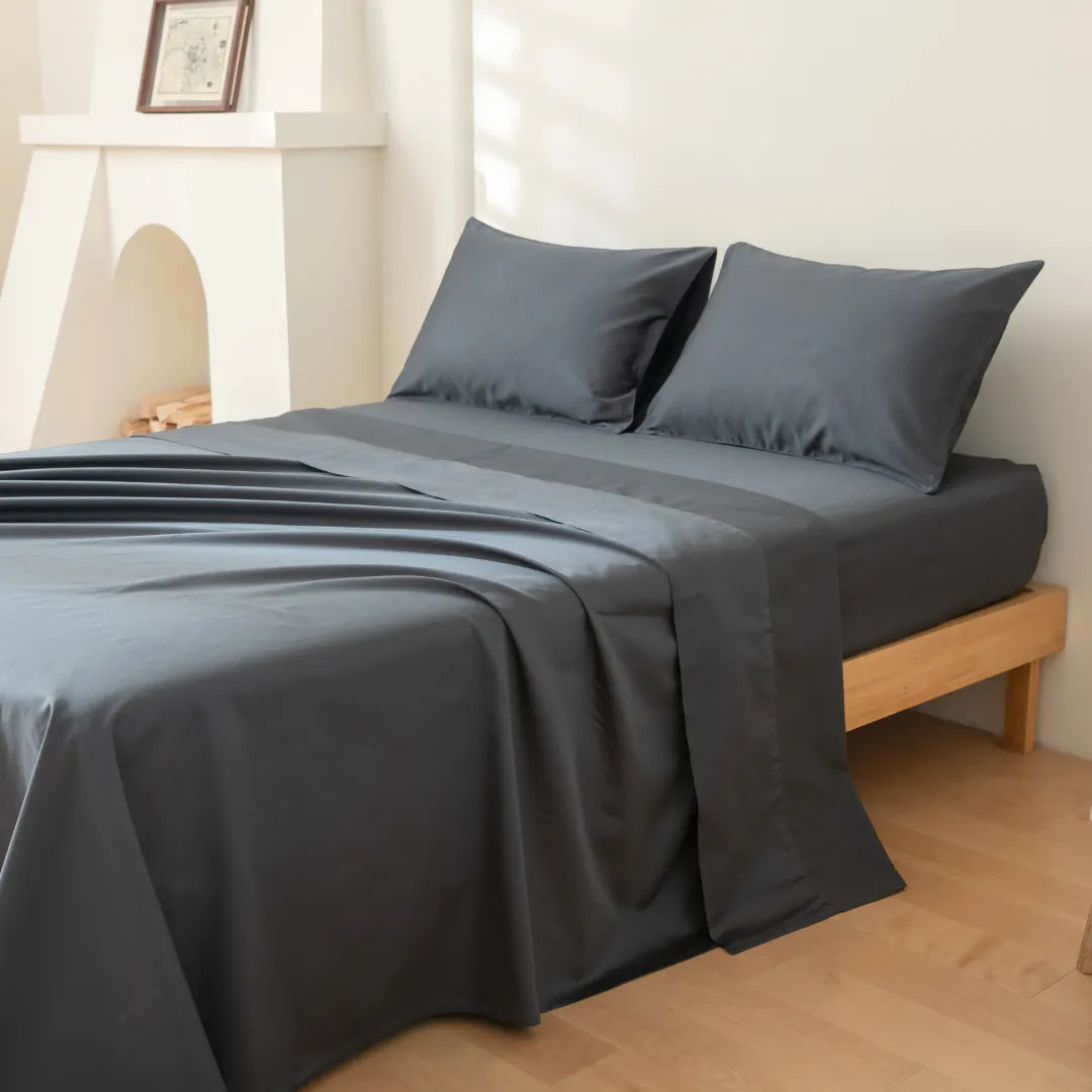 A neatly made bed with Linenly's Luxe Cotton Sateen Sheet Set in Charcoal and two matching pillows on a wooden frame sits in a bright, minimally decorated bedroom with light-colored walls and flooring.