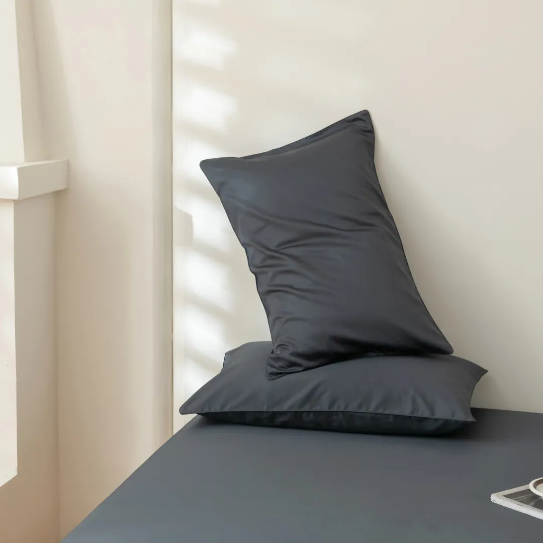 Two dark gray pillows rest atop the Linenly Luxe Cotton Sateen Sheet Set in Charcoal, with soft natural light casting shadows on a light-colored wall in the background.