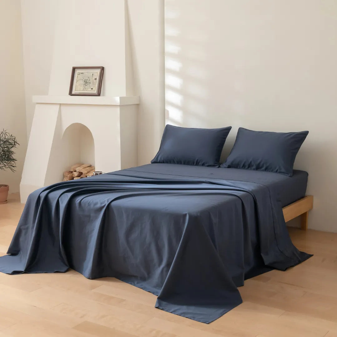 A neatly made bed with Linenly's Luxe Sateen Sheet Set in Midnight in a serene and minimalist bedroom, complemented by a small fireplace and a touch of greenery from a plant in the corner.