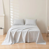 A neatly made bed with the Linenly Luxe Cotton Sateen Sheet Set in silver and two pillows stands against an off-white wall in a minimalistic room with wooden flooring and simple decor.