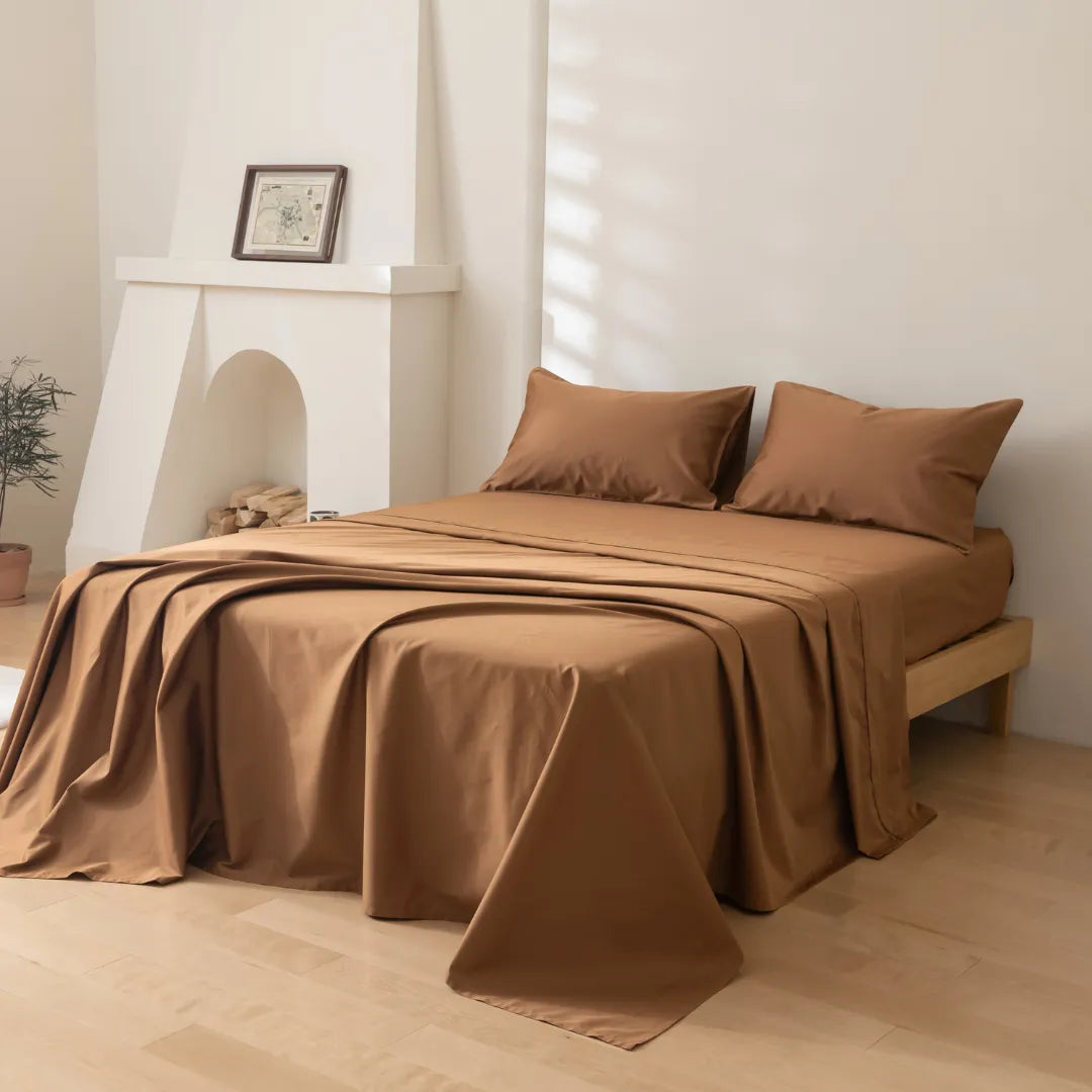 A minimal, light-filled room with wooden floors features a bed made with Linenly’s Luxe Cotton Sateen Sheet Set in Terracotta. Simple decor includes a potted plant and artwork on the white mantelpiece.
