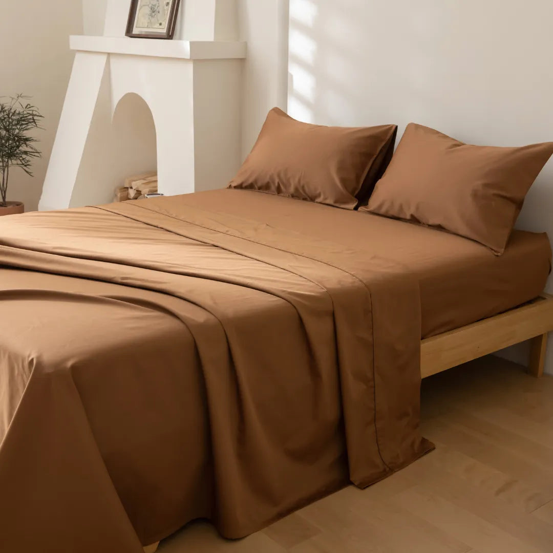 A neatly made bed with Linenly’s Luxe Cotton Sateen Sheet Set in terracotta, set in a minimal, sunlit bedroom with light wood floors, a small plant, and a white fireplace in the background.