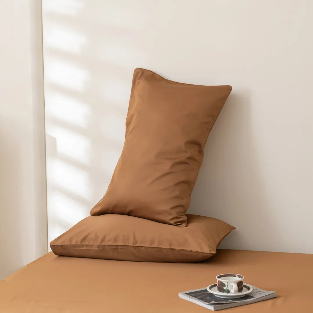 Two brown pillows rest on a matching bed made up with Linenly's Luxe Cotton Sateen Sheet Set in Terracotta; one pillow lies flat, the other upright against the wall. A cup and saucer sit on a magazine nearby as soft sunlight casts gentle shadows.