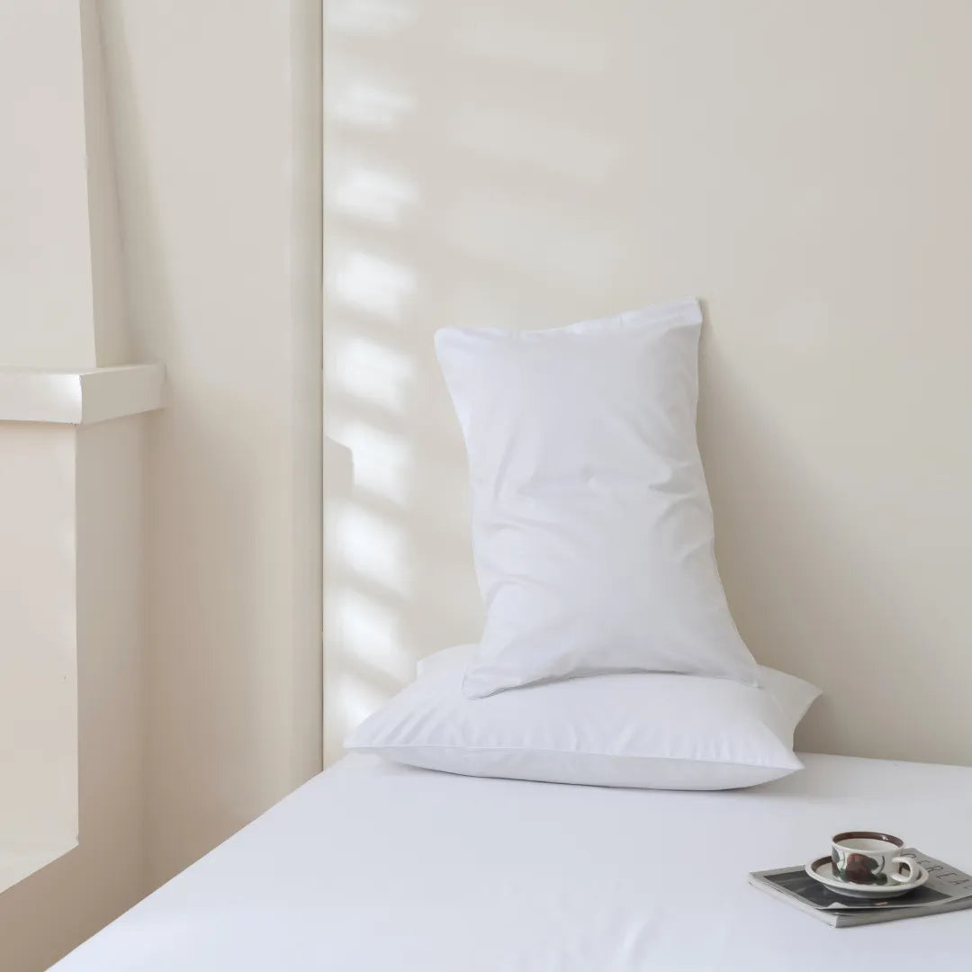 A minimal bedroom scene with two white pillows stacked on a neatly made bed in Linenly's Luxe Cotton Sateen Sheet Set - White, soft natural light streaming through blinds, and a cup of coffee with a saucer and book on the bedside.