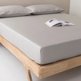 A neatly made bed with Linenly’s Bamboo Fitted Sheet Set - Silver and two pillows on a wooden frame. An open magazine lies near the pillows in this minimal, sunlit room that highlights sustainable luxury.