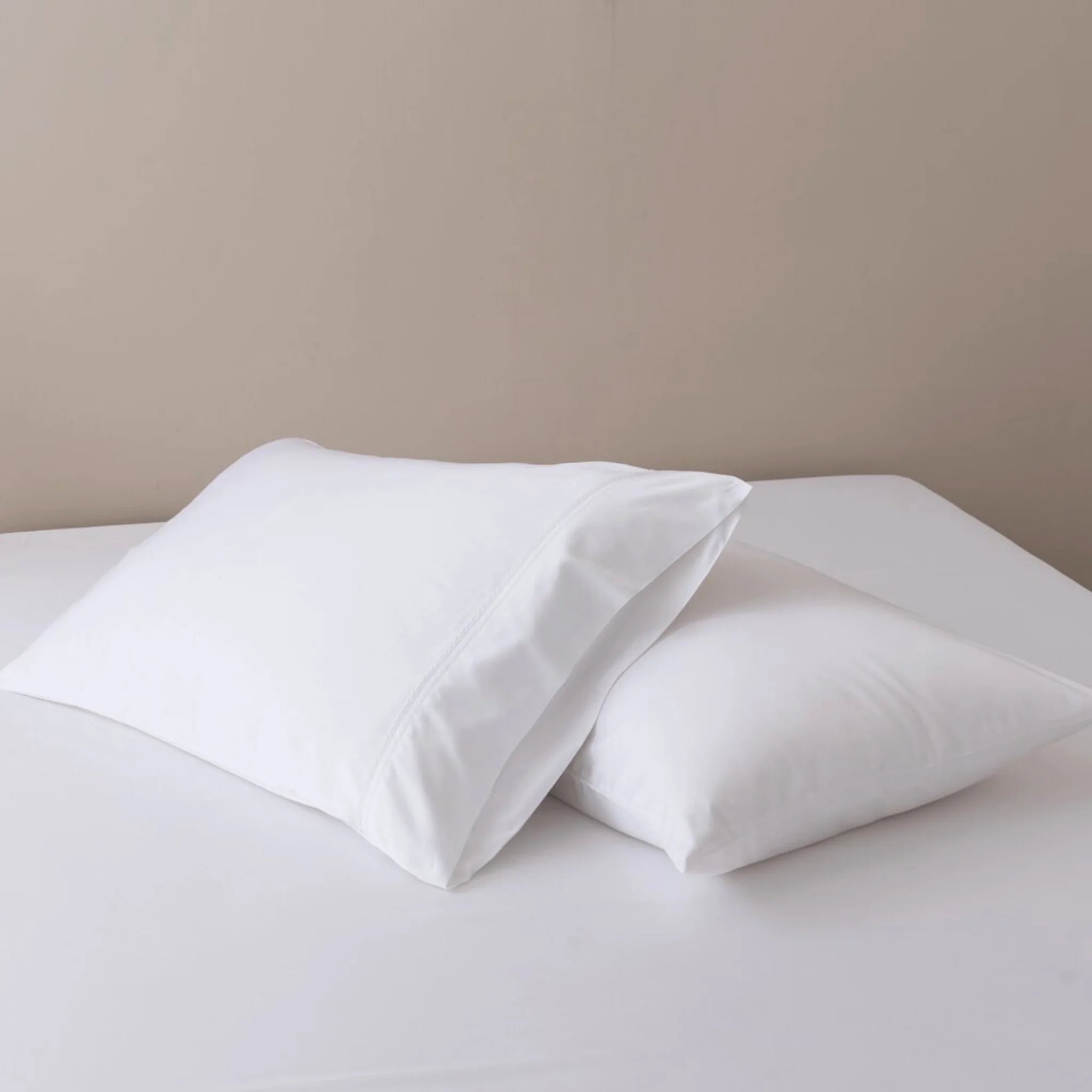 Two white pillows with Linenly Bamboo Pillowcase Set - White rest on a neatly made white bed against a plain beige wall.