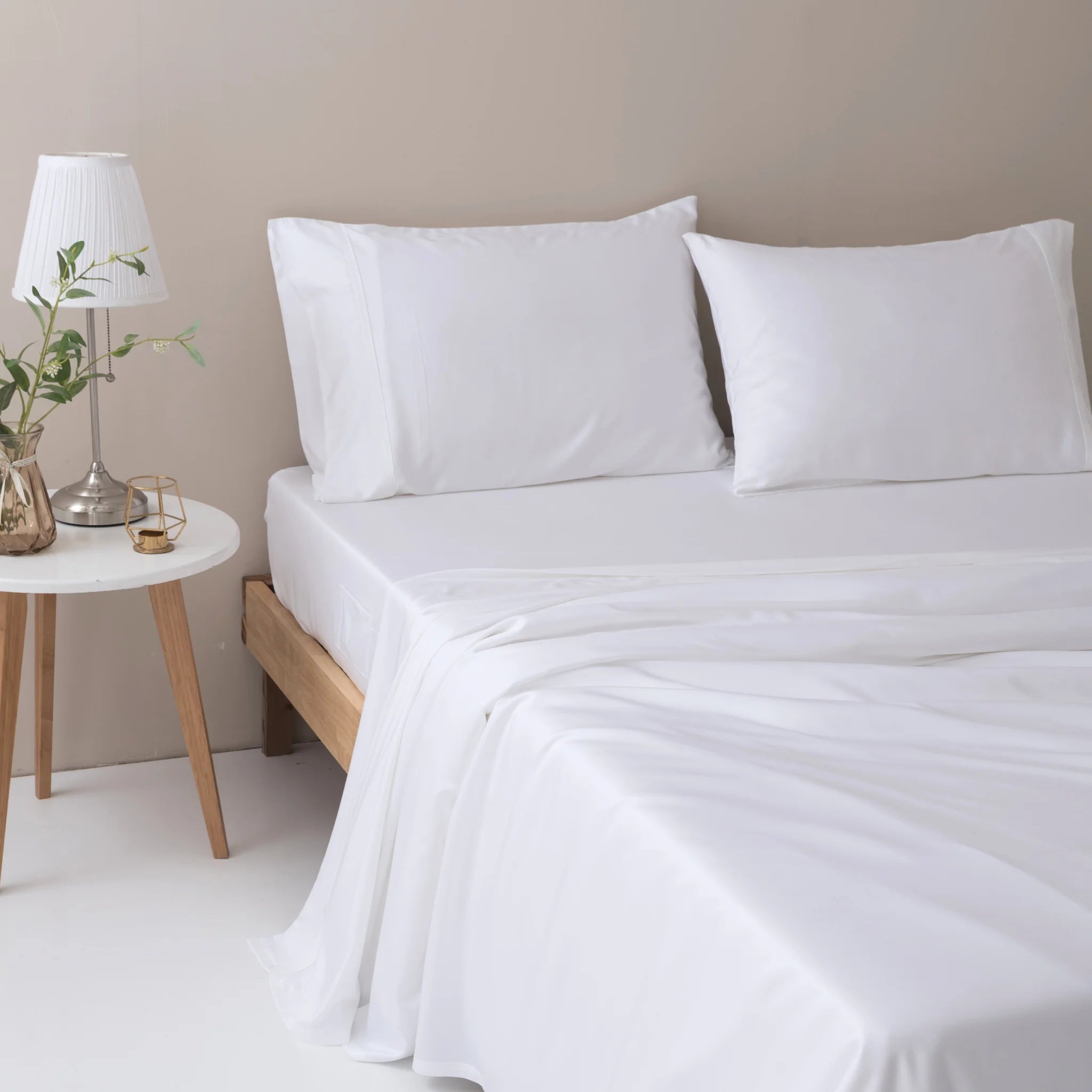 A neatly made bed features the Linenly Bamboo Pillowcase Set - White, paired with matching sheets beside a round nightstand holding a lamp, vase with green leaves, and glasses for a minimal, modern look.