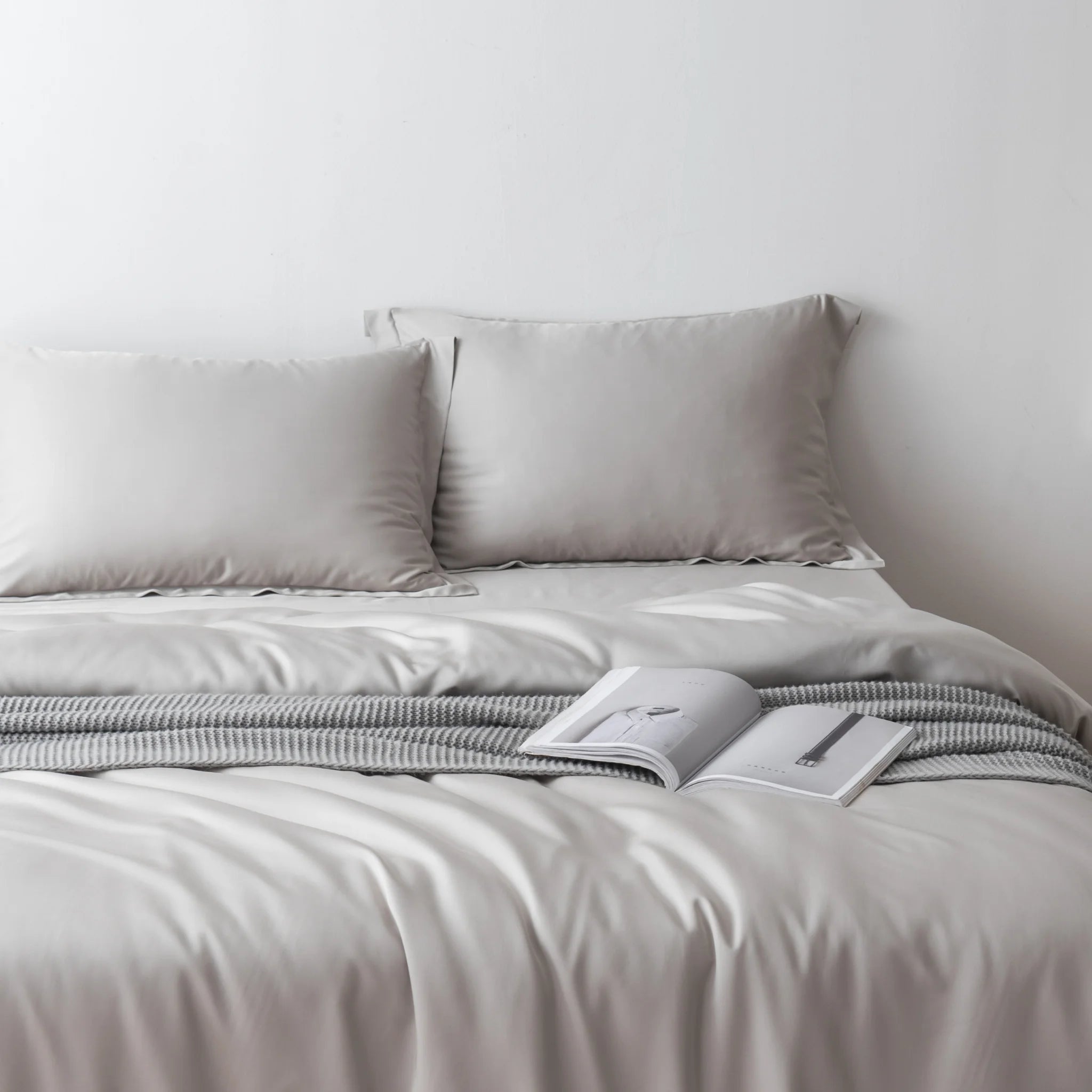A neatly made bed with the Linenly Bamboo Quilt Cover Set in Silver and two pillows is accented by a gray knitted throw and an open book, all against a plain white wall. Enjoy ultimate comfort and cooling for restful nights.