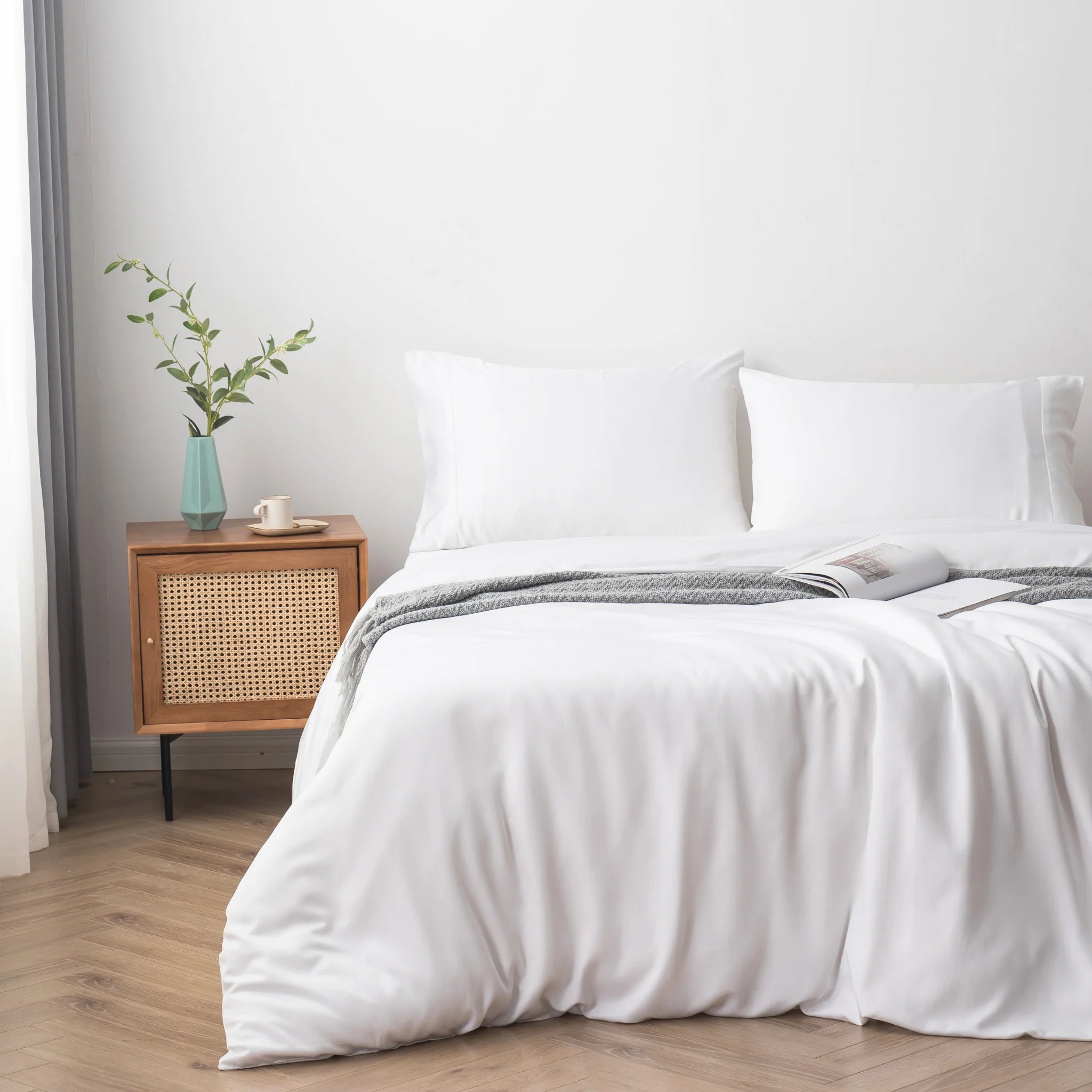 A bed made with the Linenly Bamboo Quilt Cover Set - White and two pillows sits next to a wooden nightstand holding a vase with green leaves, cup, and saucer. An open book on the bed creates a bright, minimalistic look.