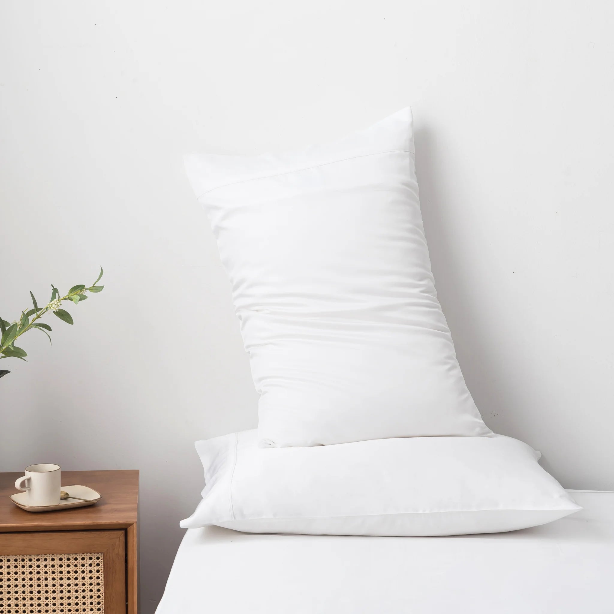 Two white pillows rest on a neatly made bed featuring the Linenly Bamboo Quilt Cover Set - White, crafted from soft bamboo materials. A wooden nightstand holds a small plant, cup, and saucer against a plain white wall.