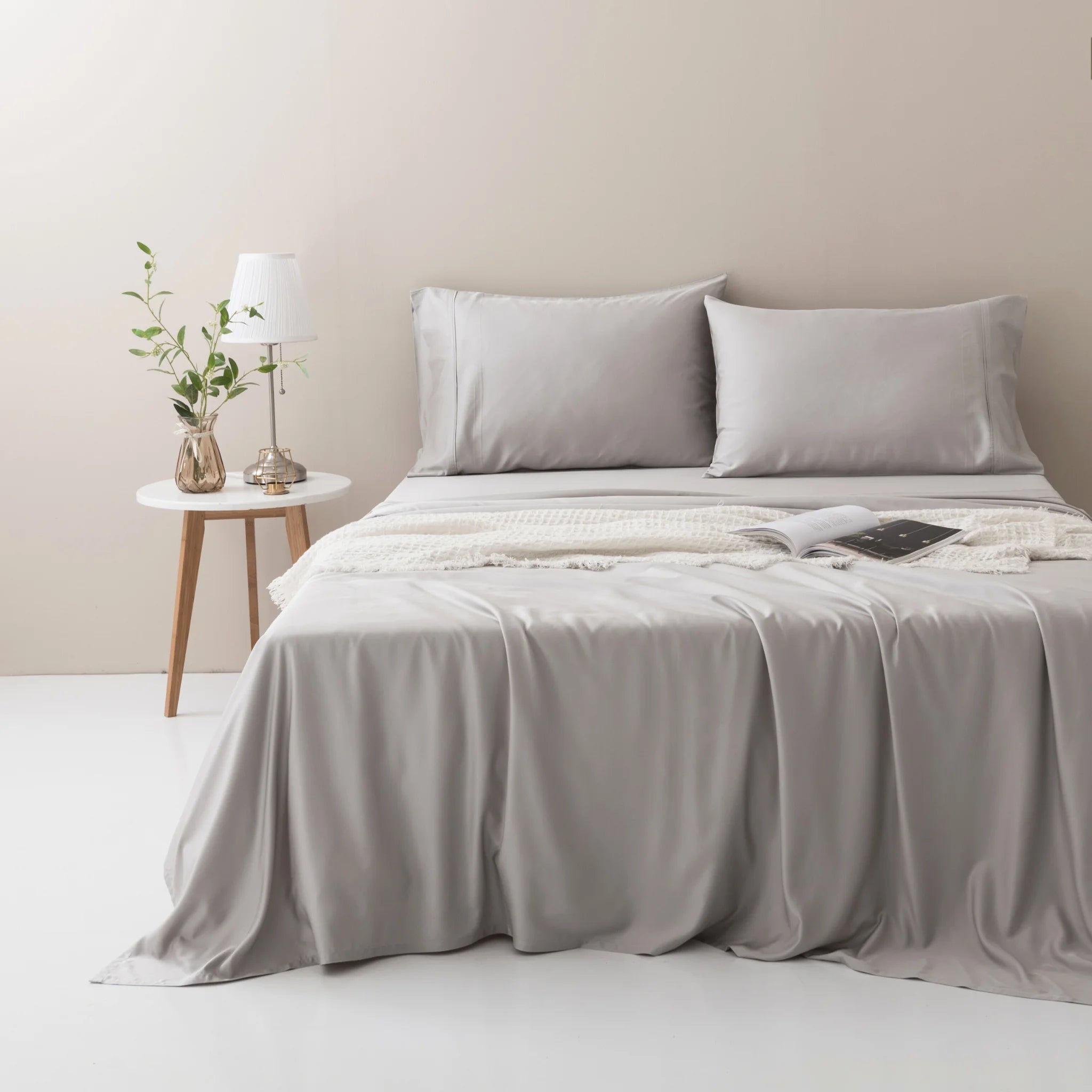 A neatly made bed features the Linenly Bamboo Sheet Set in Silver, complemented by a cream knit blanket and pillows. A small round table nearby holds a white lamp and a glass vase with green branches, creating a soft, neutral-toned space.