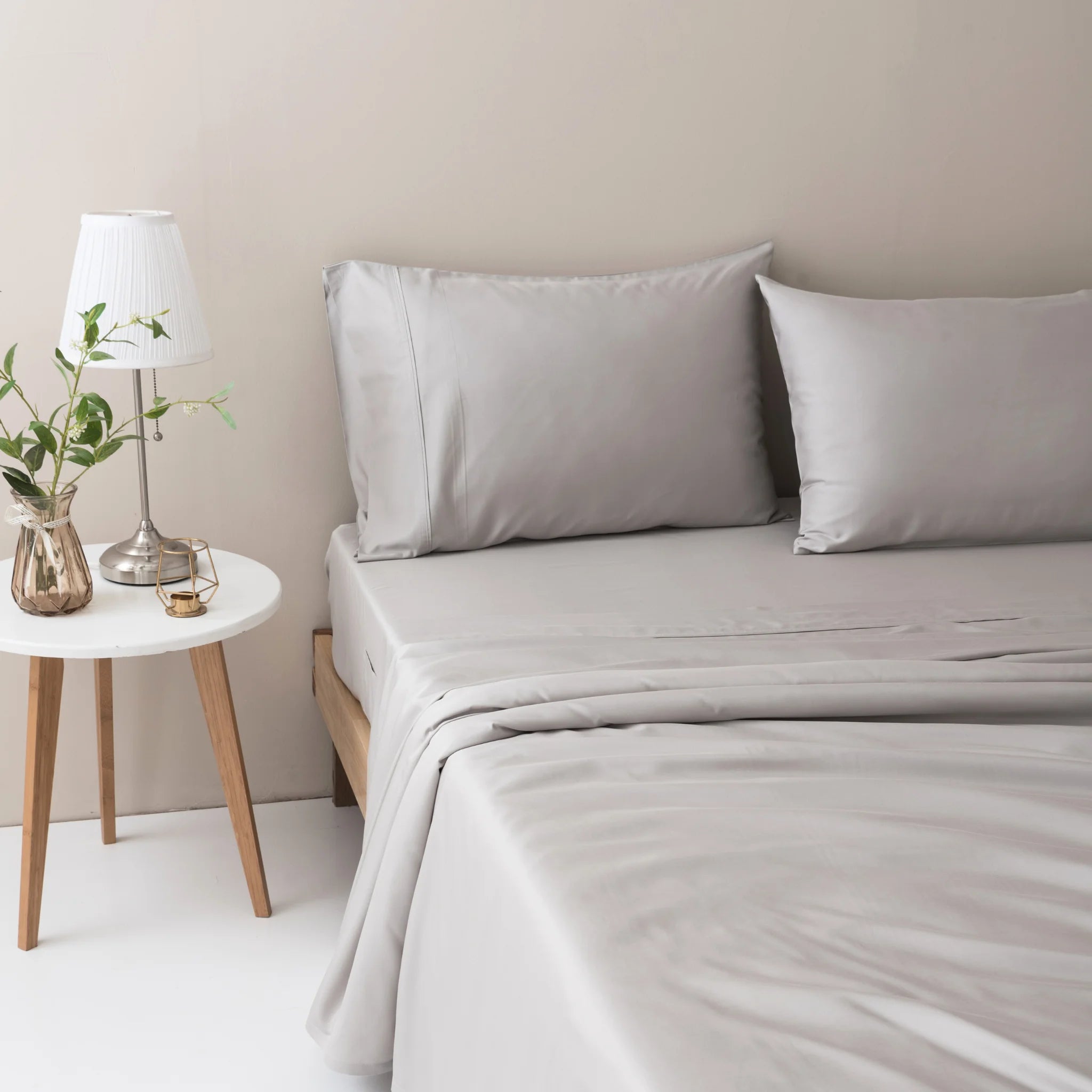 A neatly made bed features the Linenly Bamboo Sheet Set - Silver with two matching pillows, beside a round white side table holding a lamp, plant in a vase, and glass of water against a light-colored wall for a hint of luxury.